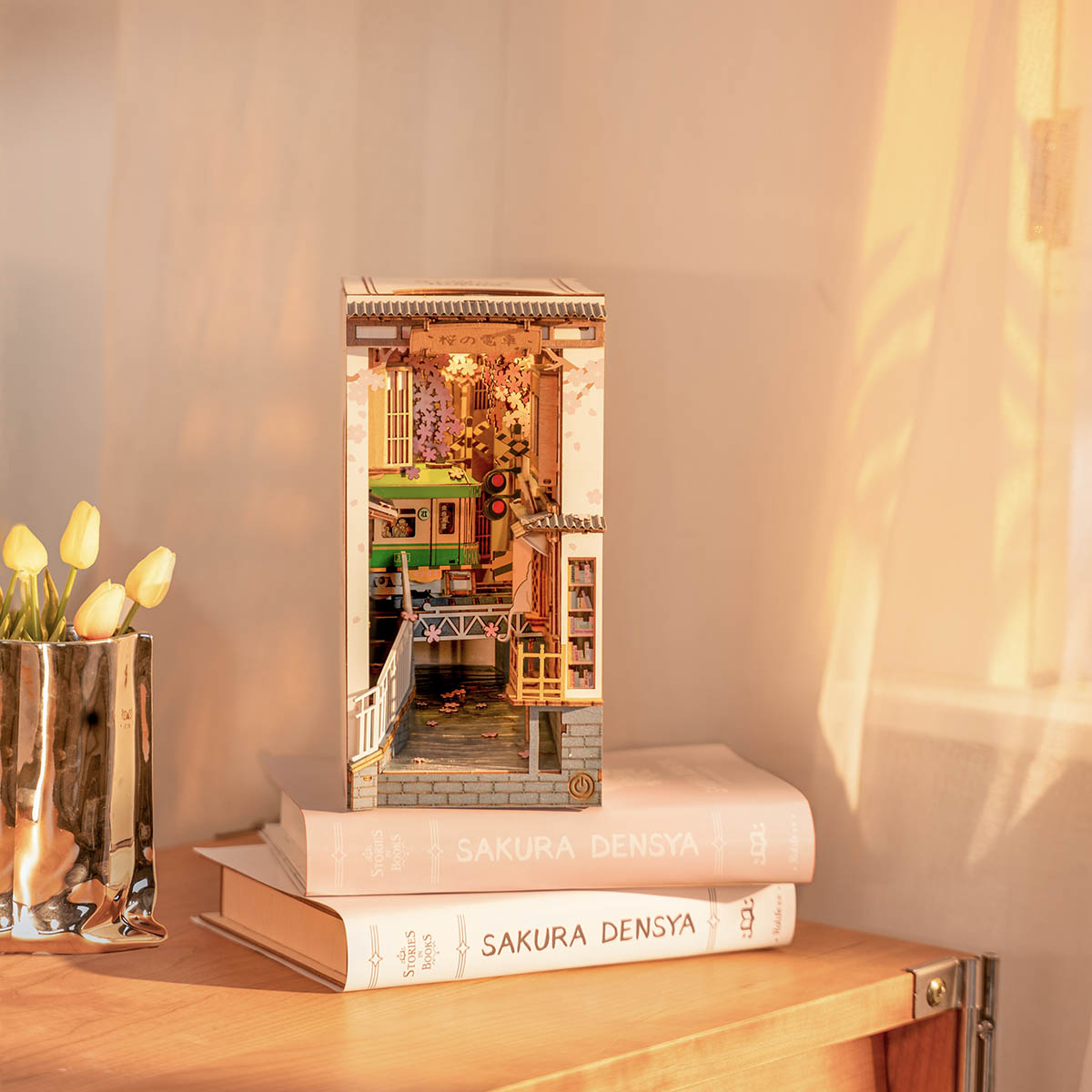 
Sakura Densya sobre libros apilados, junto a un jarrón de tulipanes y con luz de tarde.